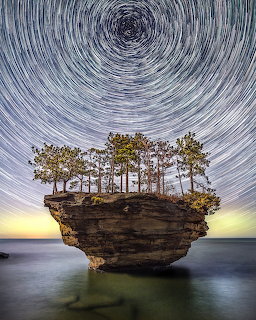 Rock island with trees in calm water under swirling star trail sky