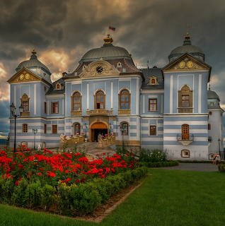 White baroque castle with multiple domes and arched windows, red flowers in foreground, stormy sky behind