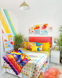 Vibrant bedroom with rainbow blanket, yellow pillows, colorful artwork, and white furniture