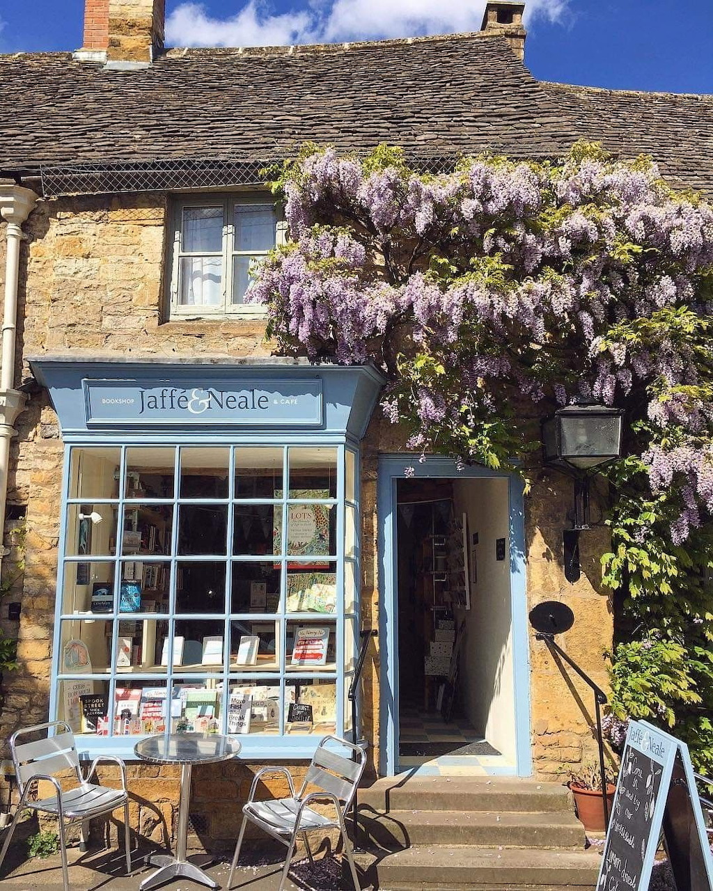 Jaffé Neale Bookshop in Cotswolds with blue shopfront, wisteria flowers, and outdoor café chairs