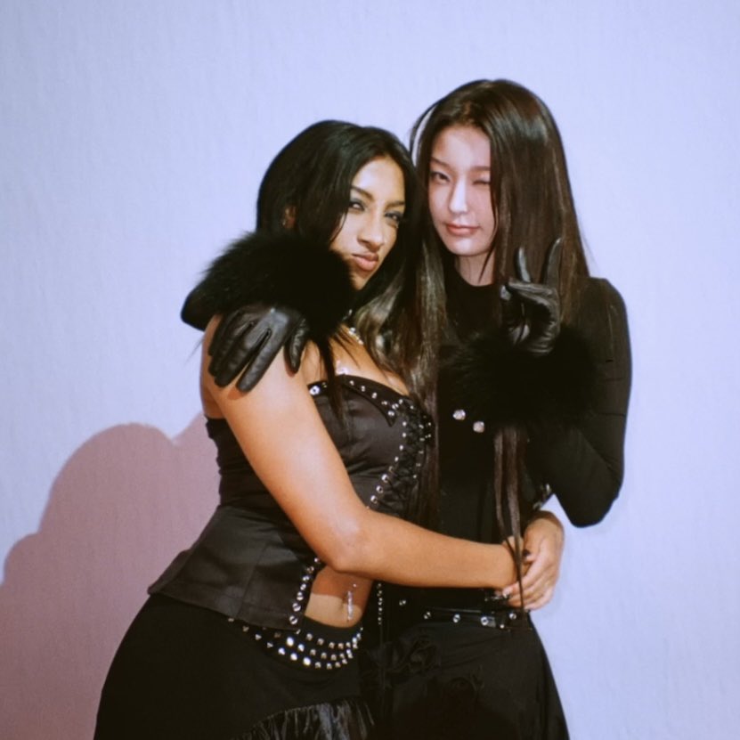 Two women in black outfits embracing and posing together against light purple wall