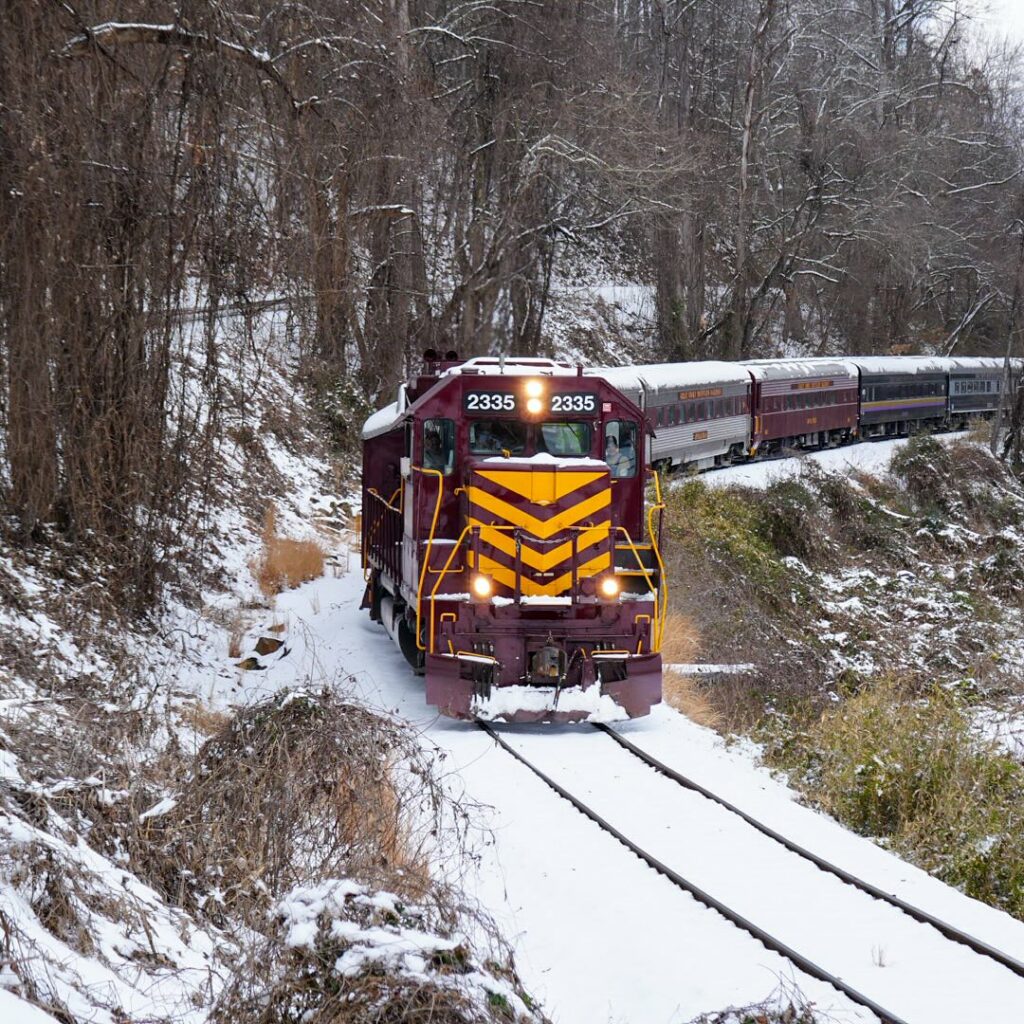 Maroon and yellow passenger train moving through snow-covered forest with bare winter trees