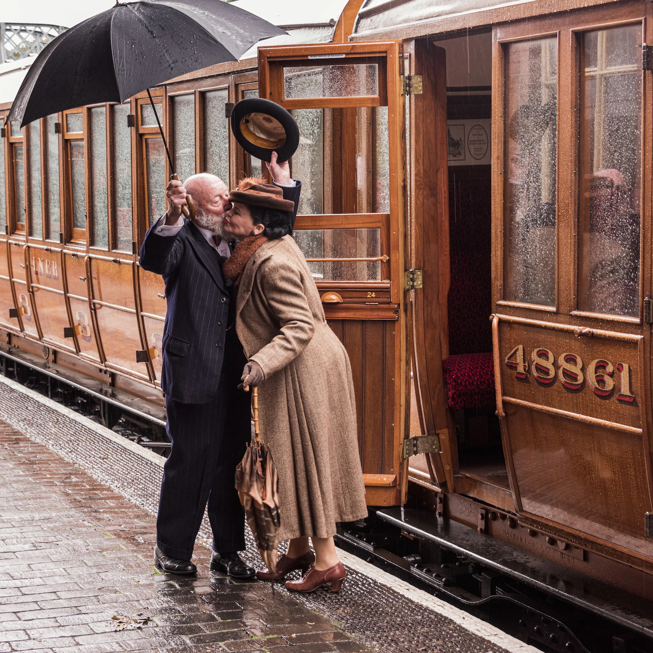 Couple with umbrella bidding farewell beside vintage wooden train carriage on rainy platform