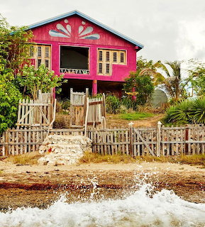 Pink painted wooden cottage on stilts with decorative roof art behind a wooden picket fence at the beach