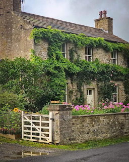 Stone cottage with green climbing vines, white garden gate, and colorful flowers in front garden