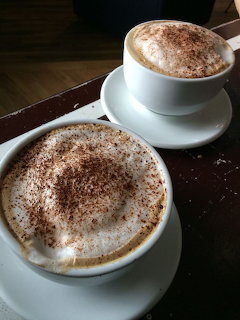 Two cappuccinos with foam and cocoa powder in white cups and saucers on a dark table