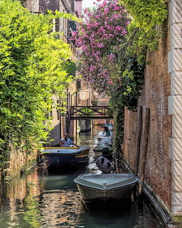Small boats in a narrow Venice canal between brick walls with lush greenery and pink flowers