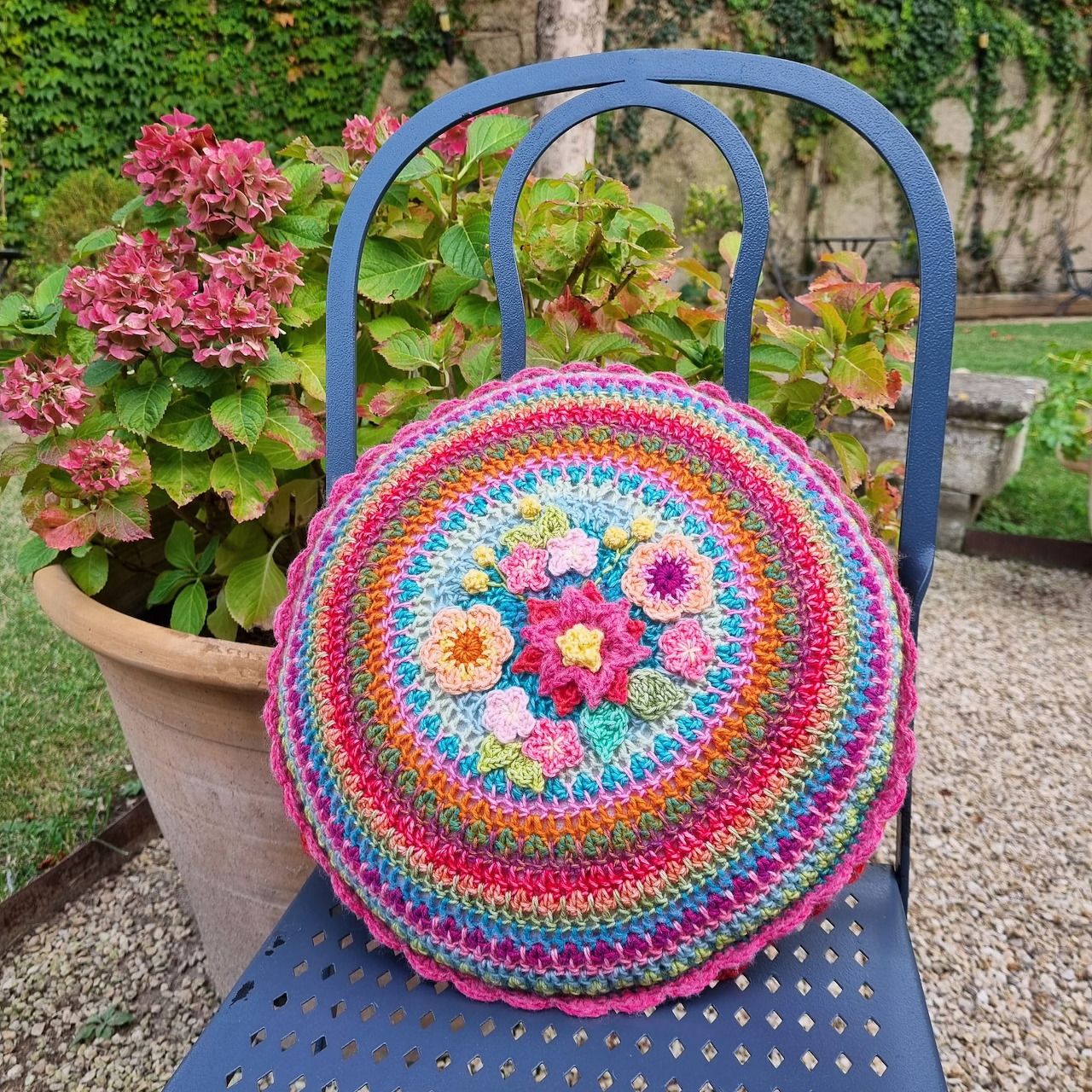 Round crochet pillow with rainbow colored rings and pink floral center on blue metal chair outdoors