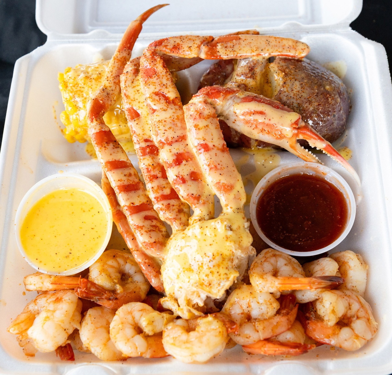 Takeout box with crab legs, shrimp, potato, corn, and two dipping sauces