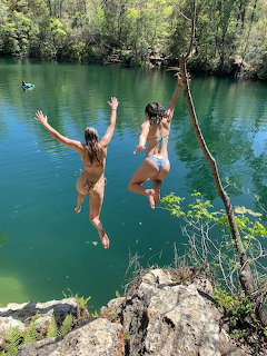 Two women mid-air jumping from rocks into a clear greenish-blue lake surrounded by trees