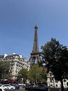 Eiffel Tower above Paris street with trees, buildings, and cars under a blue sky