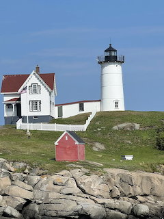 White lighthouse and keeper's house on grassy hill above rocky shore with blue sky