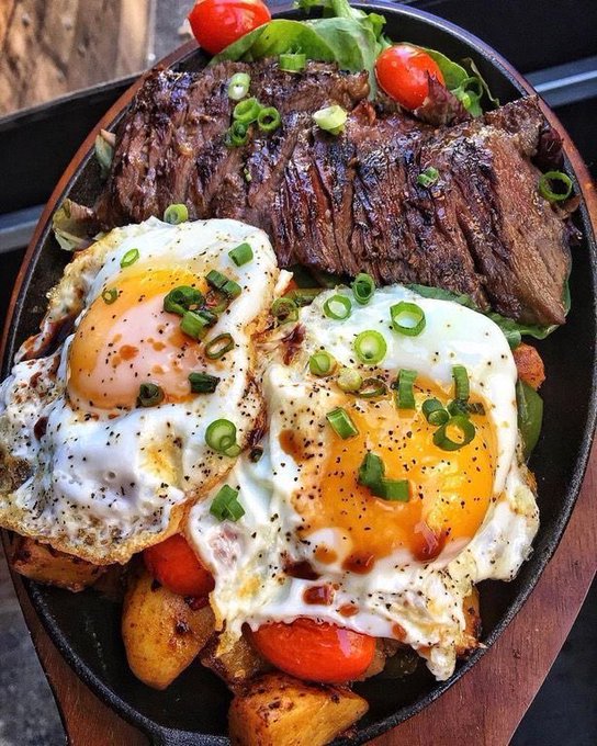 Cast iron skillet with grilled steak, two sunny-side-up eggs, roasted potatoes, tomatoes, and green onions