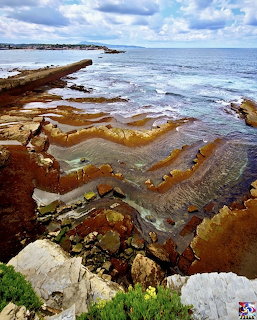 Rocky coastline with layered orange and brown sandstone formations and ocean waves under cloudy sky