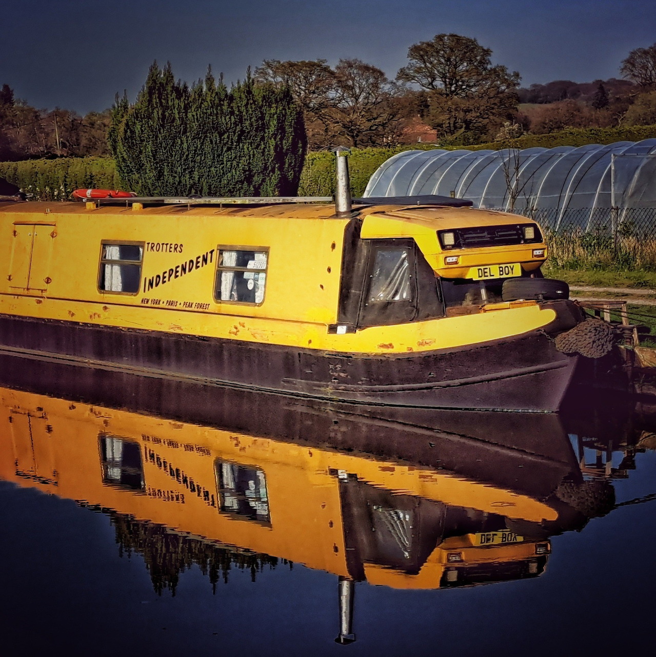 Yellow narrowboat moored on still canal water creating mirror reflection with trees in background