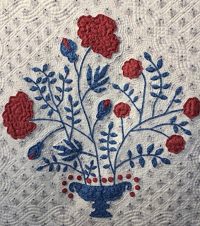 Quilted fabric art with red flowers and blue foliage in vase on white background with diagonal stitching
