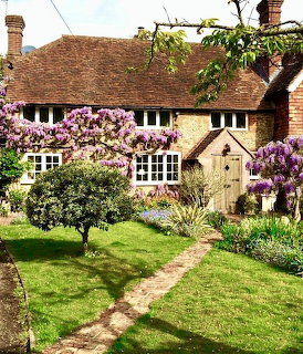 Stone cottage with purple wisteria blooms, green lawn, and garden path leading to arched entrance
