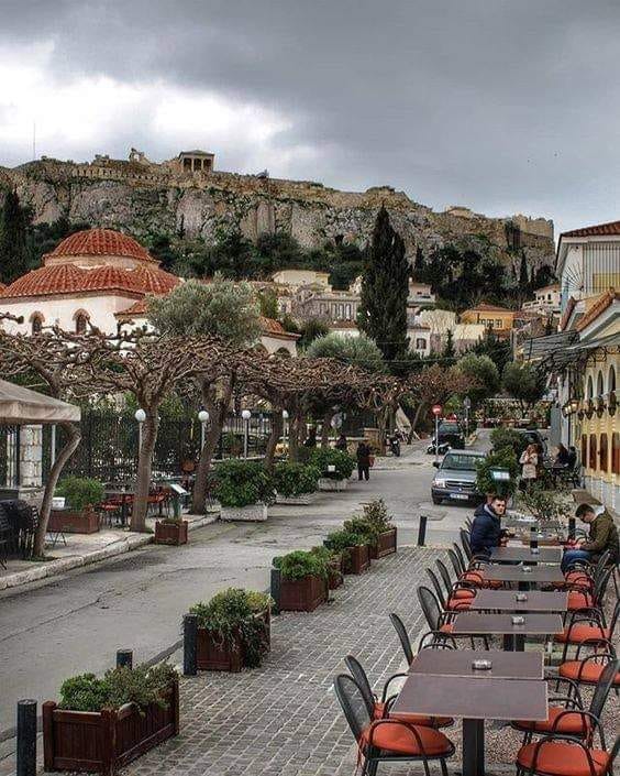 Athens street scene with Acropolis hill and ancient ruins visible above cafes and buildings under cloudy sky
