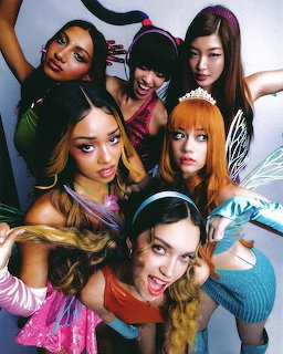 Seven young women in colorful outfits posing together with playful expressions and fairy wing accessories