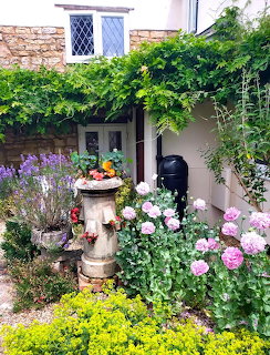 Cottage garden entrance with climbing vines, roses, lavender, and stone fountain surrounded by colorful flowering plants