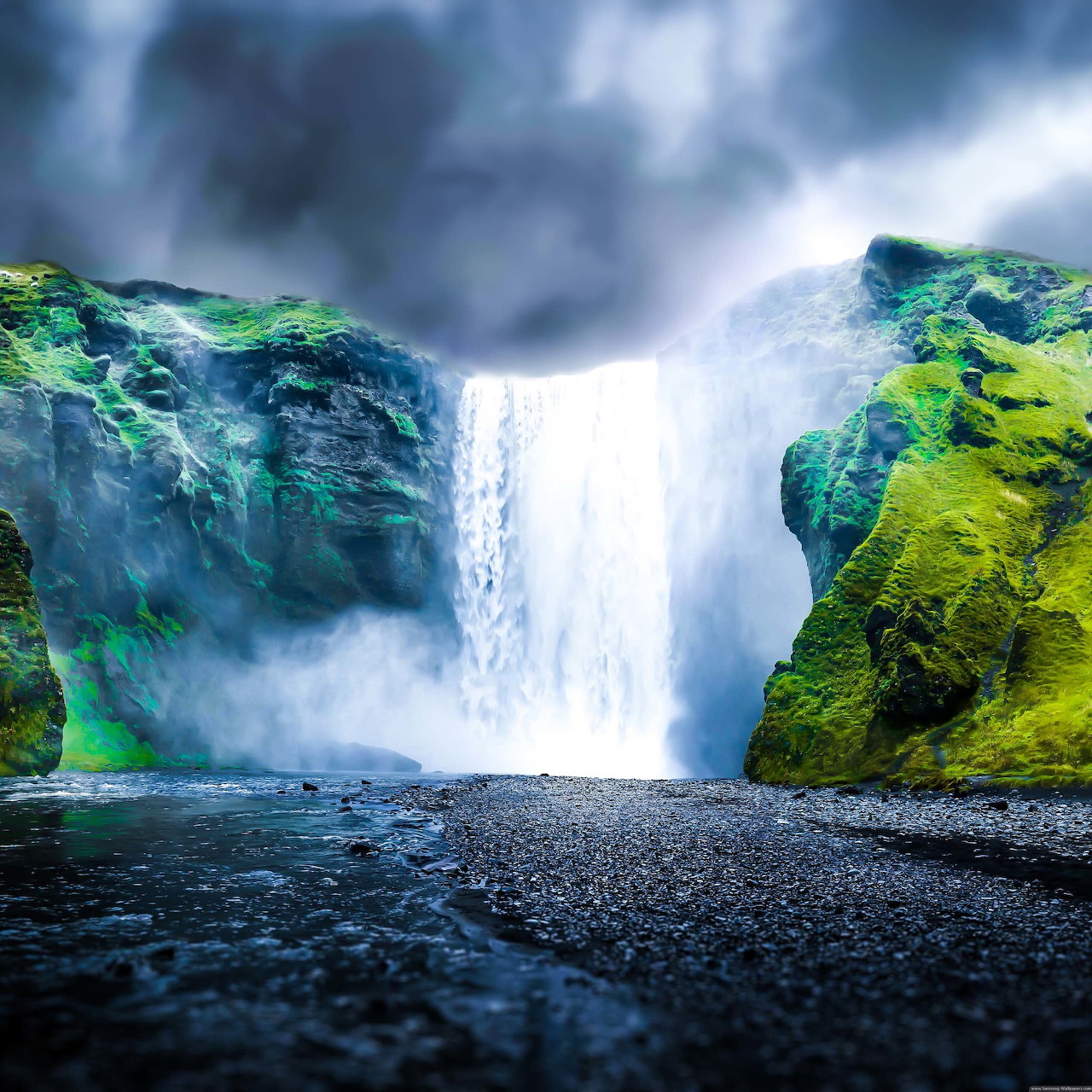 Powerful waterfall between green mossy cliffs with stormy sky and dark rocky beach