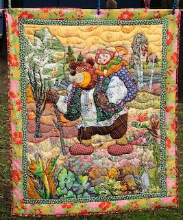 Quilted wall hanging showing Santa Claus carrying toys in a winter forest with patchwork floral border