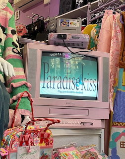Pink CRT TV showing Paradise Kiss anime title screen surrounded by colorful vintage items and clothing