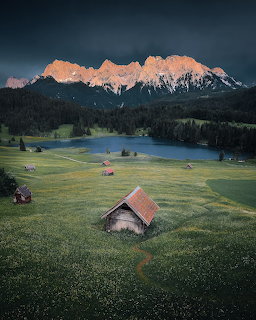 Wooden hut in an alpine meadow with a lake, forest, and sunlit mountain peaks behind