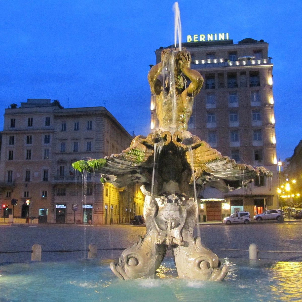 Triton Fountain illuminated at dusk in a city square with surrounding historic buildings