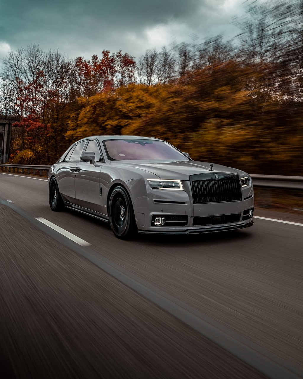 Grey Rolls-Royce luxury car driving on mountain road with moody sky and autumn landscape