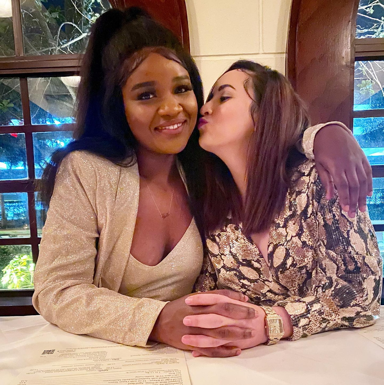 Two smiling women embracing, one kissing the other's cheek in an indoor setting