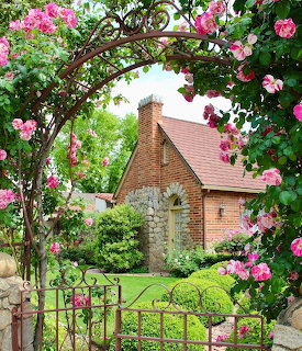 Brick cottage with stone doorway framed by pink rose arbor in green garden