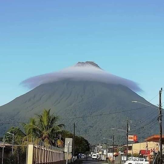 Volcano with a cloud cap rising behind a small town and palm trees under a blue sky