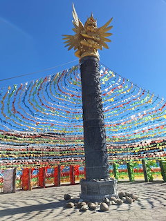 Tall pilar with golden top surrounded by radiating colorful Tibetan prayer flags under blue sky