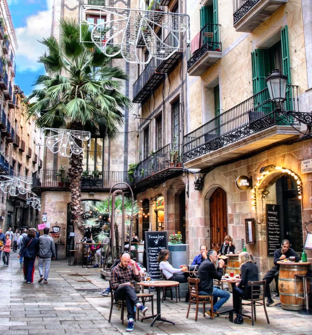 People dining at outdoor cafés on a narrow European street with palm trees and ornate balconies