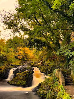 Waterfall flowing over rocks beneath green and autumn-colored trees along a stone pathway