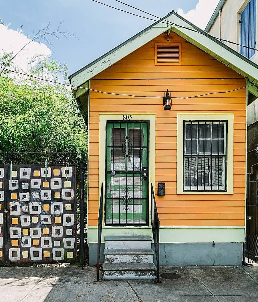Small yellow-orange house front with green trim, steps, barred door and windows, and a patterned fence