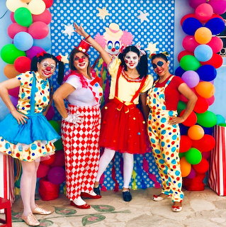 Four women in colorful clown costumes posing by a balloon arch and blue polka dot backdrop