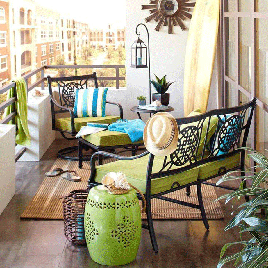 Sunny balcony with black furniture, green and blue cushions, plants, and city buildings in background