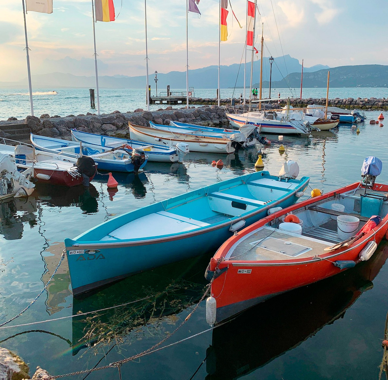 Colorful boats including blue and red vessels moored at a calm marina dock with mountains in background