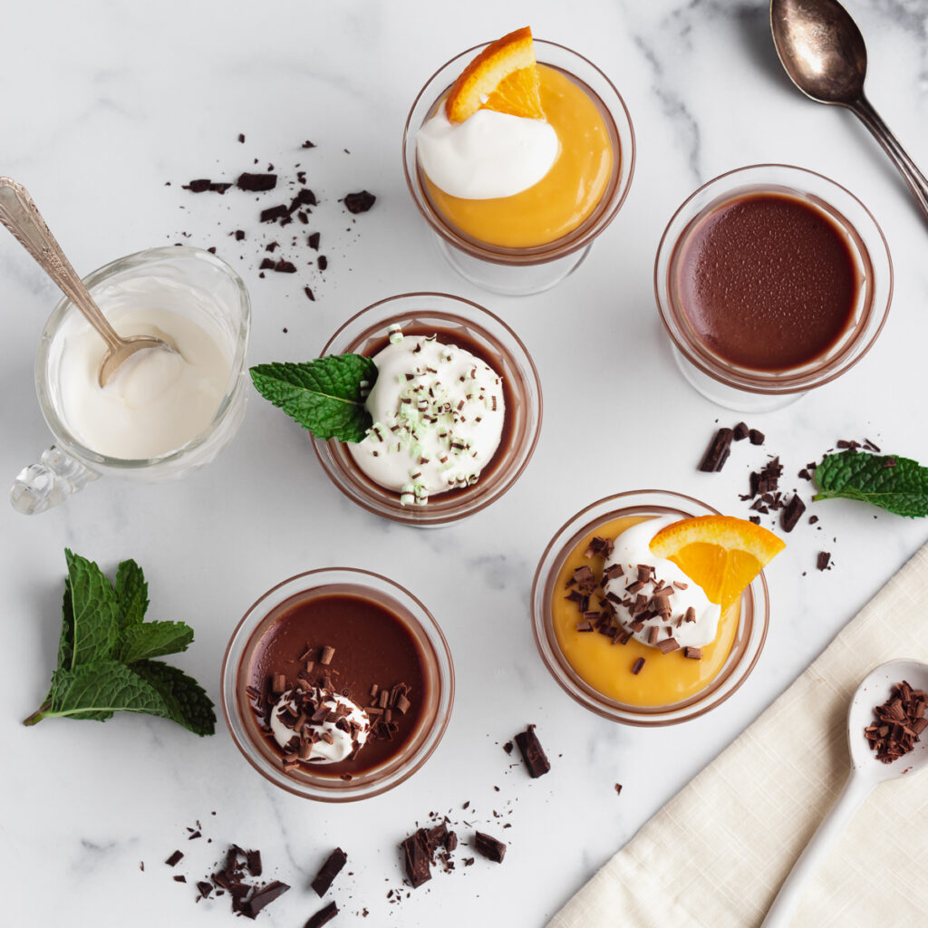 Assorted panna cotta desserts in glass cups with chocolate, vanilla, and orange flavors, topped with mint and chocolate