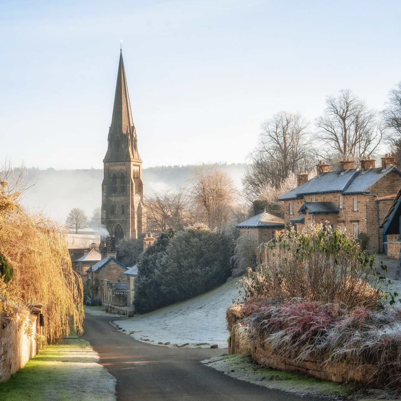 Church spire above a frosty village road at sunrise with mist and bare trees