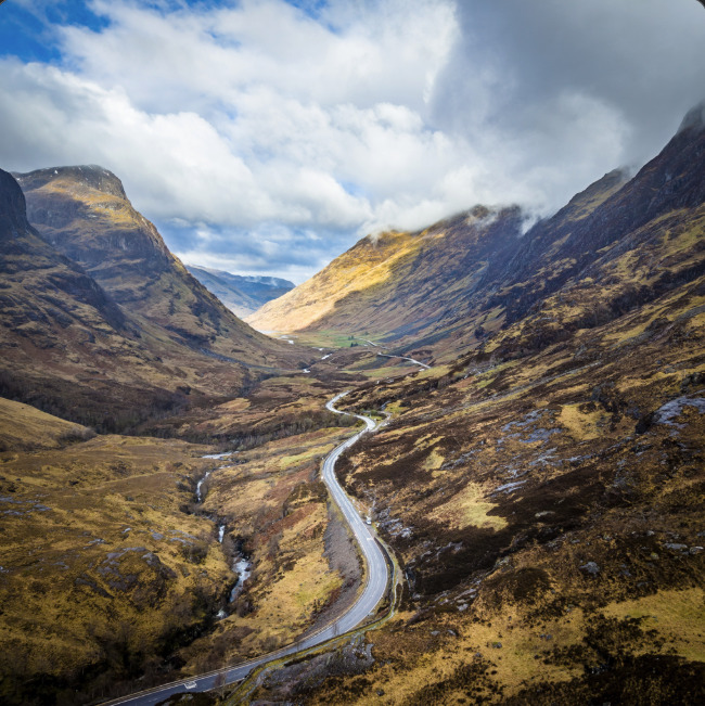 Winding road through Glen Coe valley in Scotland with rugged mountains and cloudy sky
