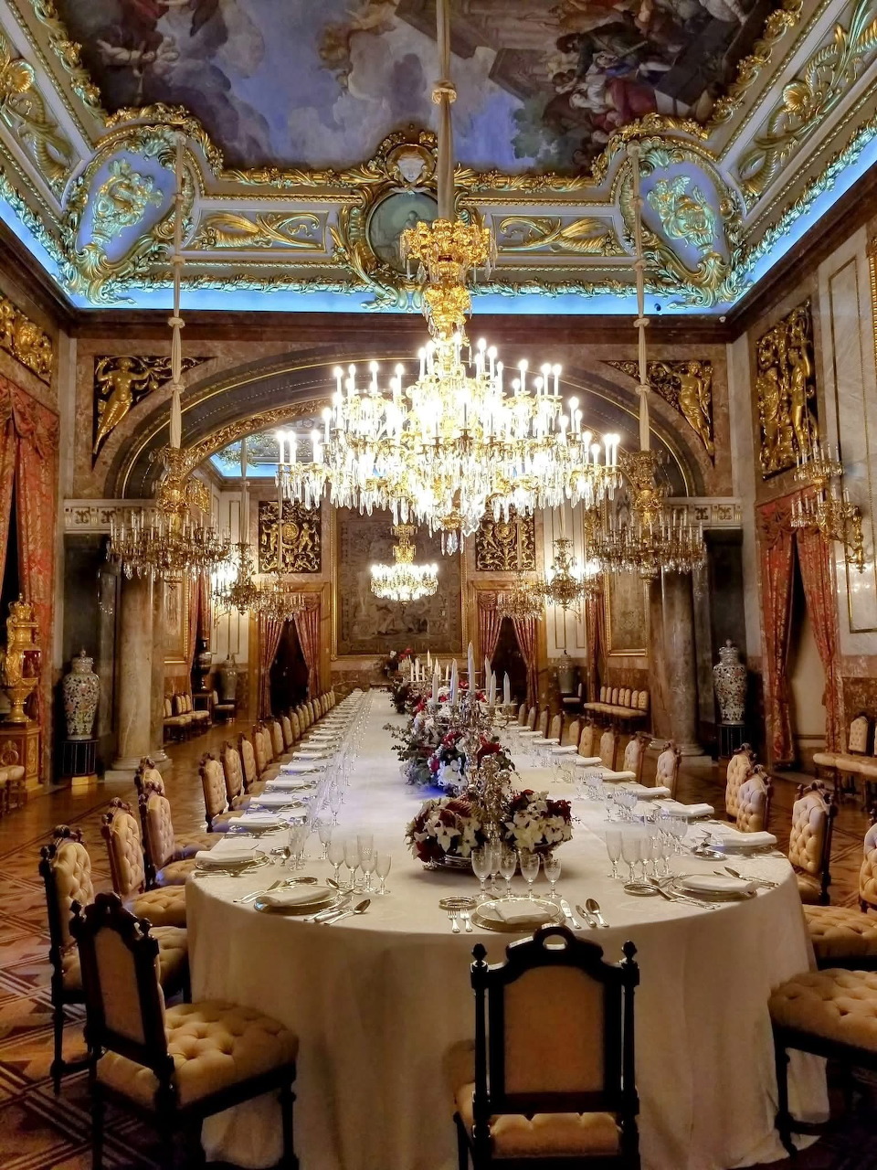 Lavish palace dining room with crystal chandeliers, ornate golden ceiling, mirrors, and long formal banquet table