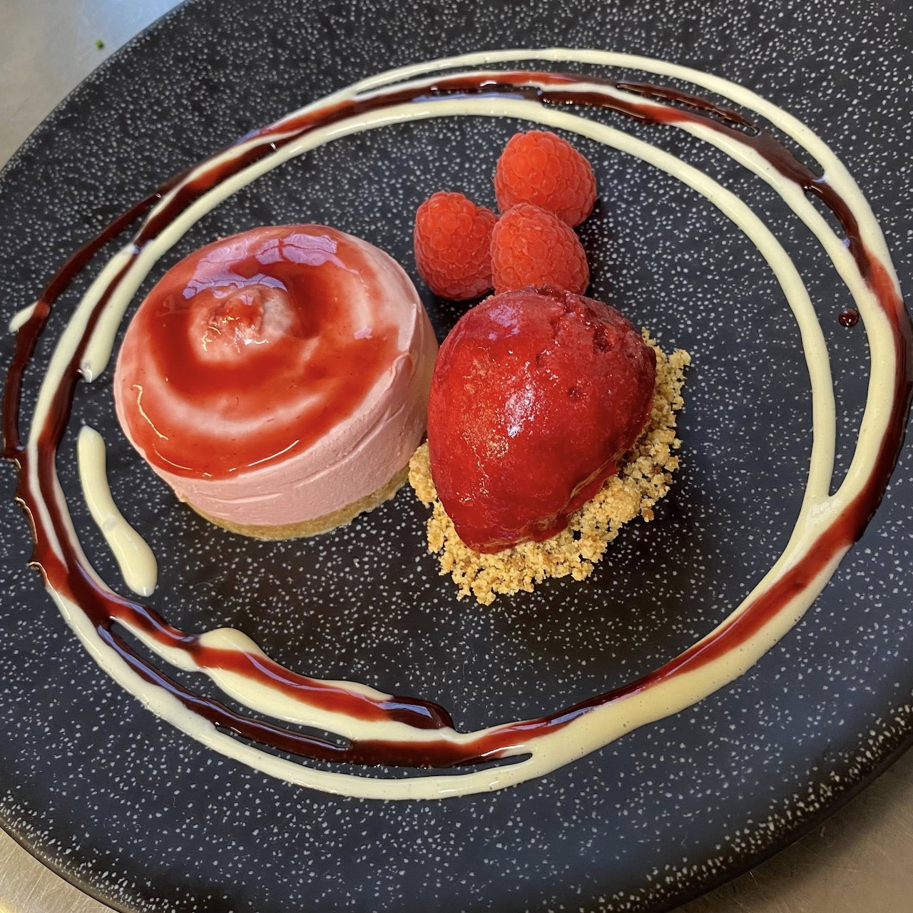 Plated raspberry dessert with mousse, glazed pastry, fresh berries and sauce on dark plate