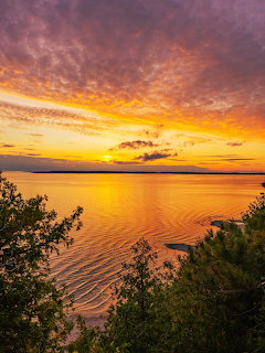 Dramatic sunset with orange and purple clouds reflecting on calm water, silhouetted trees and rocks in foreground