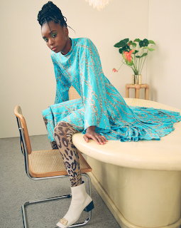 Woman in a blue patterned dress sitting on the edge of a cream bathtub indoors