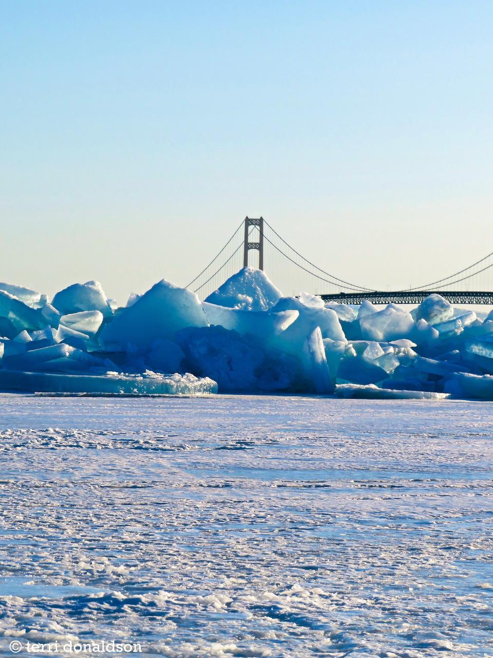 Blue ice chunks piled on frozen lake shore with suspension bridge in background under clear sky
