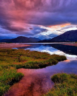Colorful sunset with pink and purple clouds reflected in a calm loch with green marshland and mountains