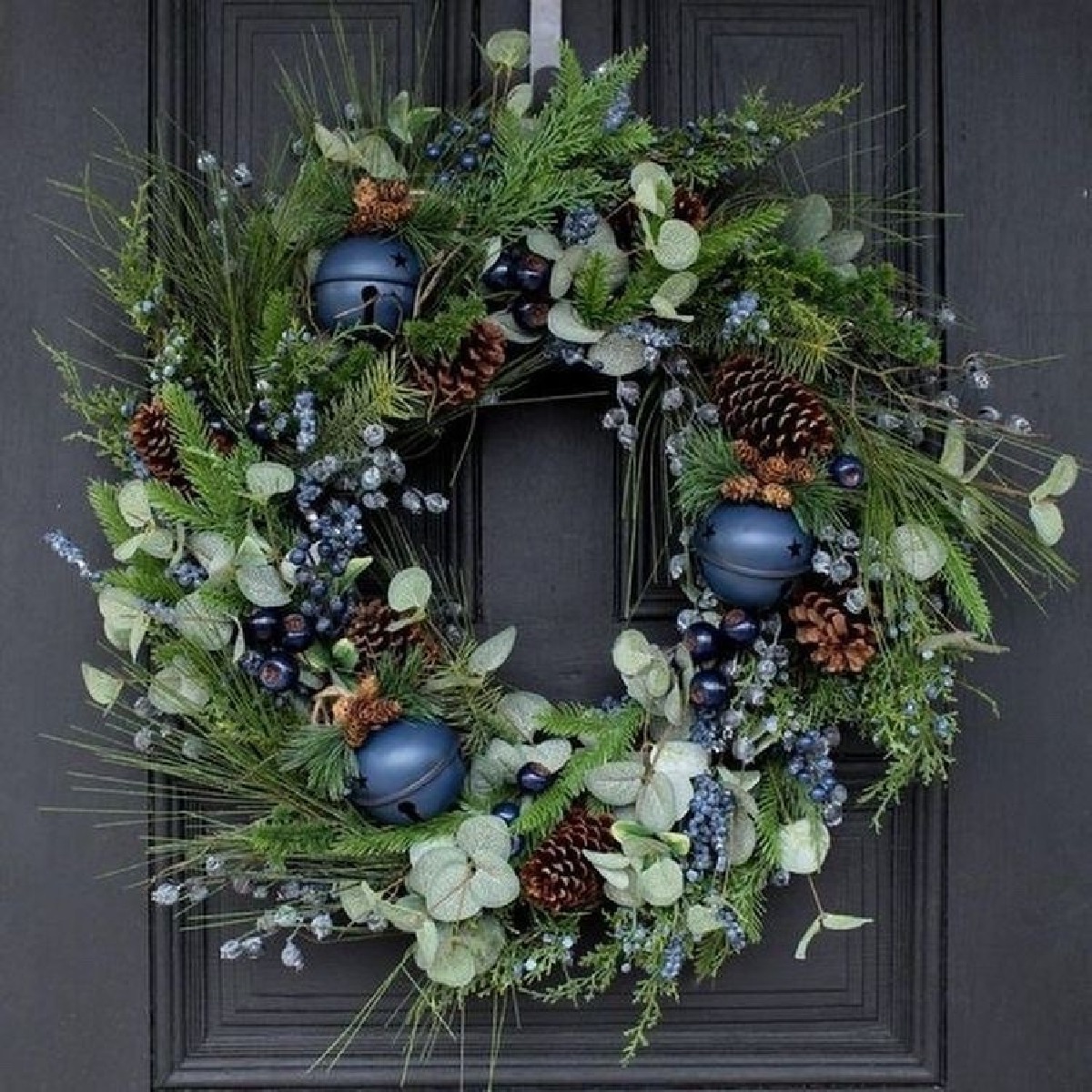 Festive green wreath with pinecones, berries, pale leaves, and blue ornaments on a dark door